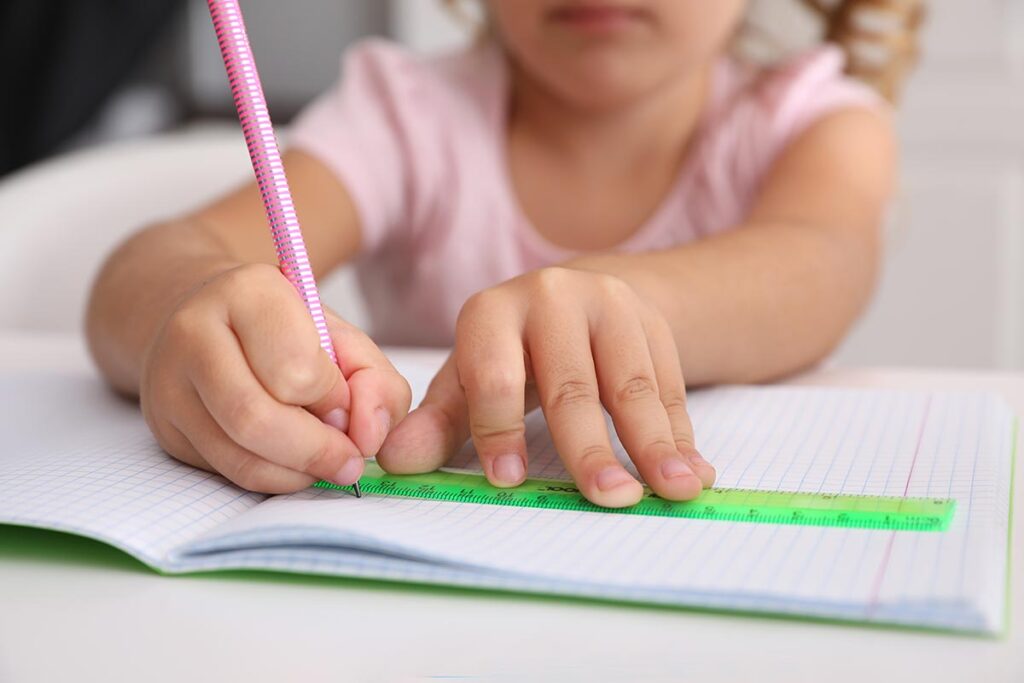 A young girl sits at a table, focused on writing in her notebook with a pencil, surrounded by colorful stationery.