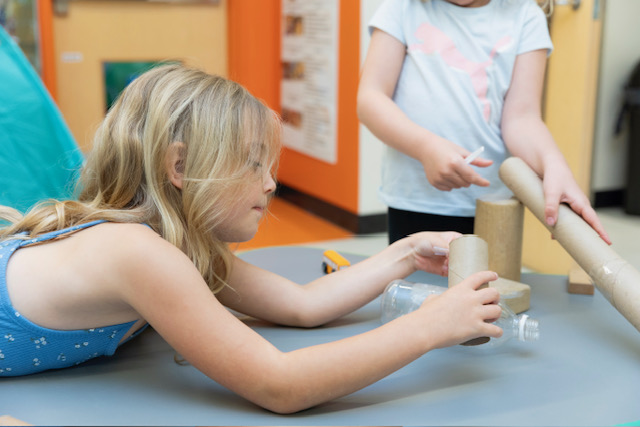 Two children are building a structure using cardboard tubes, a plastic bottle, and wooden blocks on a table in a classroom.