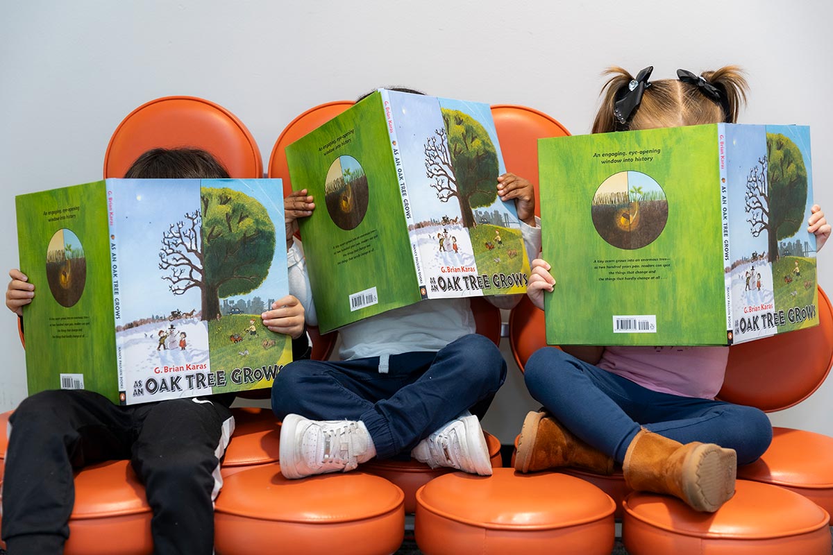 Three children sit on orange chairs, each holding and reading a copy of the book "As an Oak Tree Grows," with their faces mostly hidden behind the covers.