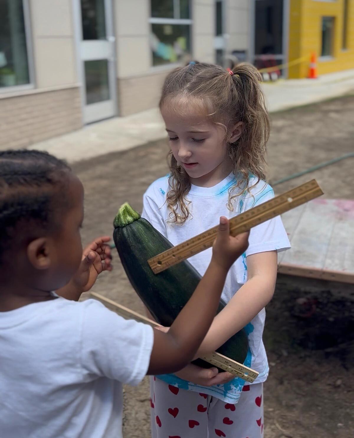 Two children proudly hold a large cucumber in front of a building, smiling and enjoying their harvest.